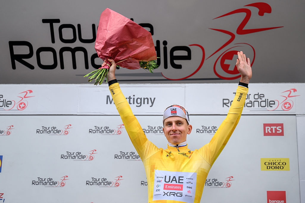 Tadej Pogačar stands on the podium at the Tour de Romandie, wearing the yellow jersey as race leader, with his arms raised in celebration holding a bouquet