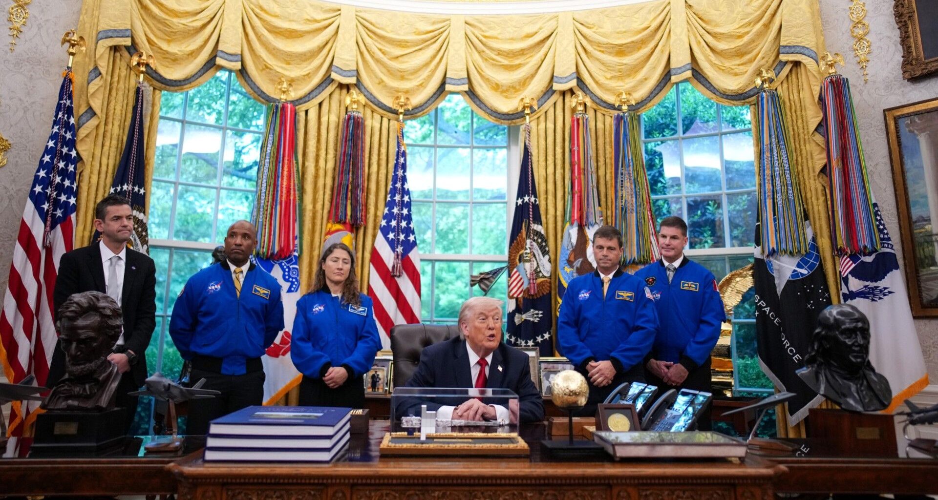 four people in blue flight jackets stand behind an old man at a desk in a grotesquely decorated office