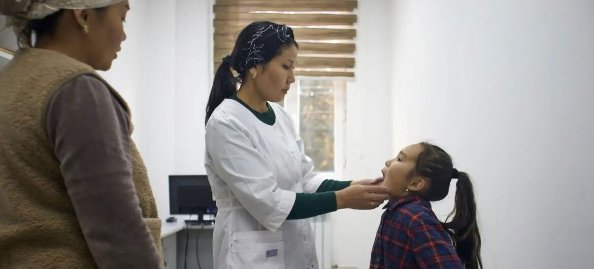 A female doctor in a white coat examines a young girl's throat while a woman looks on in a medical clinic.