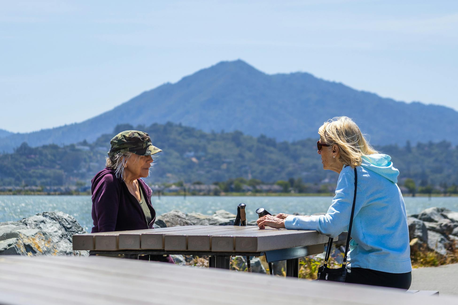 Two senior women sitting by a lake, enjoying conversation with scenic mountain views.