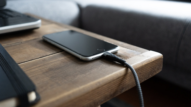Smartphone plugged into a charger and resting on a nightstand by a bed.