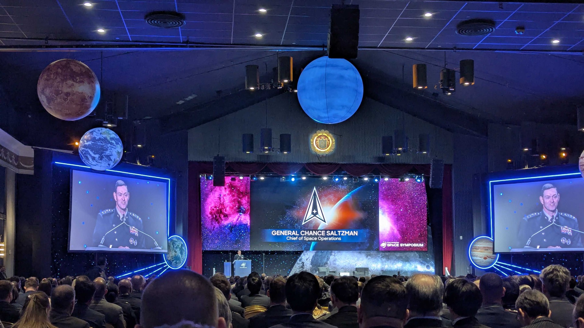 a man in a black military uniform gives a speech on a stage decorated with planets and stars in a large ballroom full of seated attendees