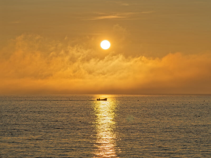 A small boat floats on calm water during a golden sunrise or sunset, with the sun low in the sky and its reflection shimmering on the surface amid orange clouds and mist.