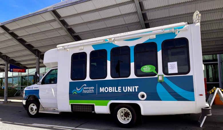 A five window, small white bus being used as a Nova Scotia Health mobile unit is parked outside an emergency department.