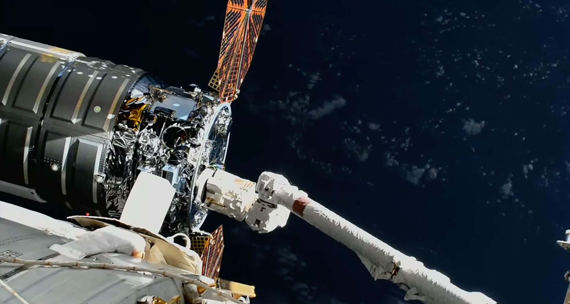 A large robotic arm reaches and grapples a silver-cylindric spacecraft with round solar arrays outside a space station.
