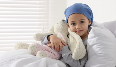 Child with head scarf holds a stuffed rabbit in a hospital room bed.