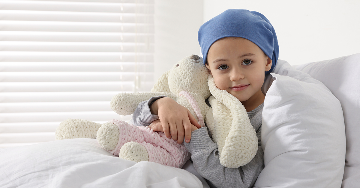 Child with head scarf holds a stuffed rabbit in a hospital room bed.
