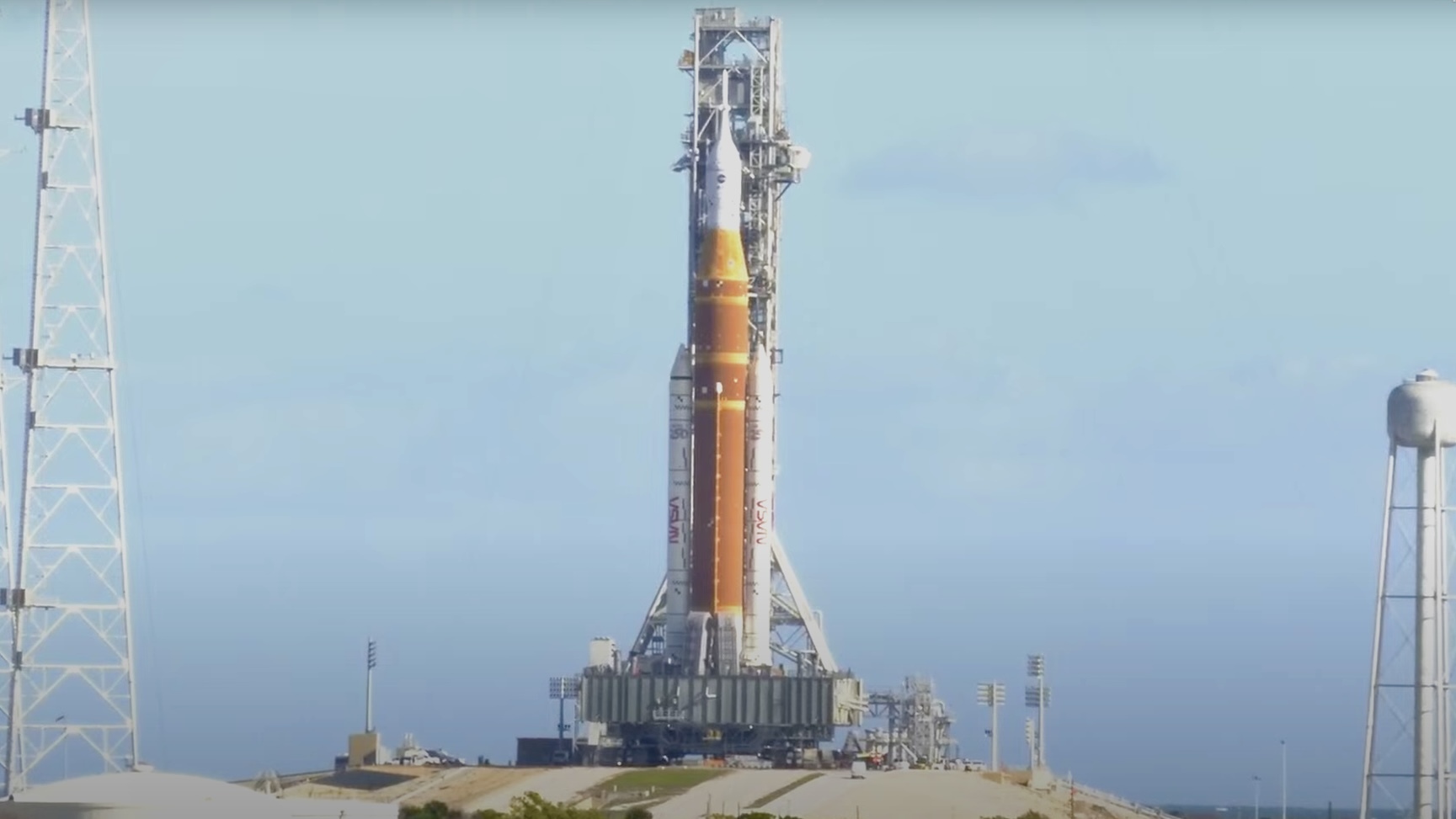 An orange rocket stands on a mound before a pale blue sky.