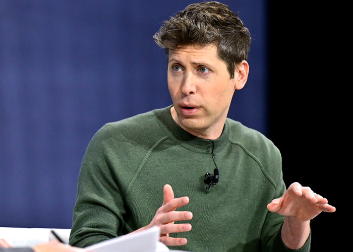 Sam Altman speaking in a green sweater, gesturing with hands during a public discussion on stage.