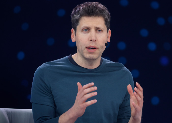 OpenAI CEO Sam Altman speaking with headset microphone, wearing a blue shirt against a dark background.