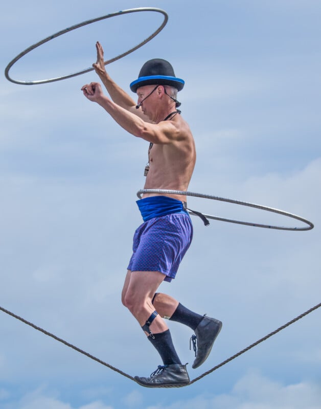 A man wearing a bowler hat, blue shorts, and black socks balances on a tightrope while spinning two hula hoops—one around his waist and one in his hand—against a blue sky.