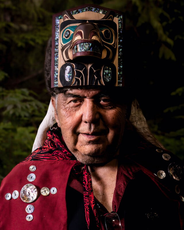 An elder wearing a traditional robe adorned with decorative buttons and a carved, painted wooden headdress stands in front of a blurred, leafy background.