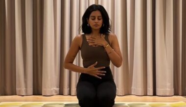 Woman kneels on yoga mat with her eyes closed, one hand on her sternum and her other hand on her stomach