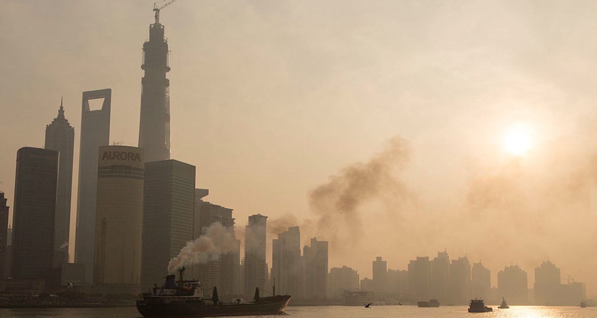 A smog-filled cityscape on the edge of a body of water is seen under a clouded over sun