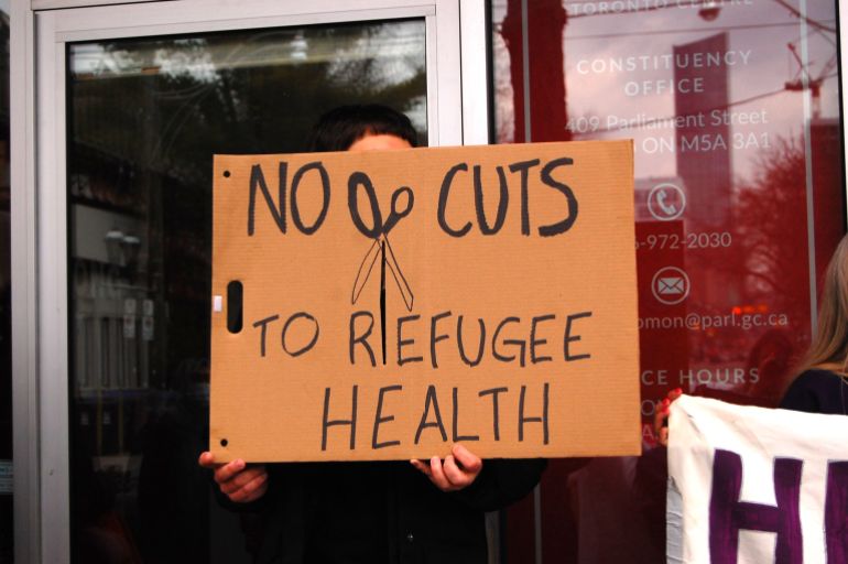 A protester holds a sign that reads, 'No cuts to refugee health'