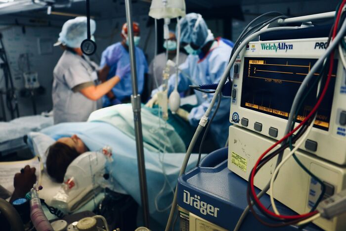 Hospital staff in protective gear attending to patient in an emergency room with medical monitors and equipment nearby.