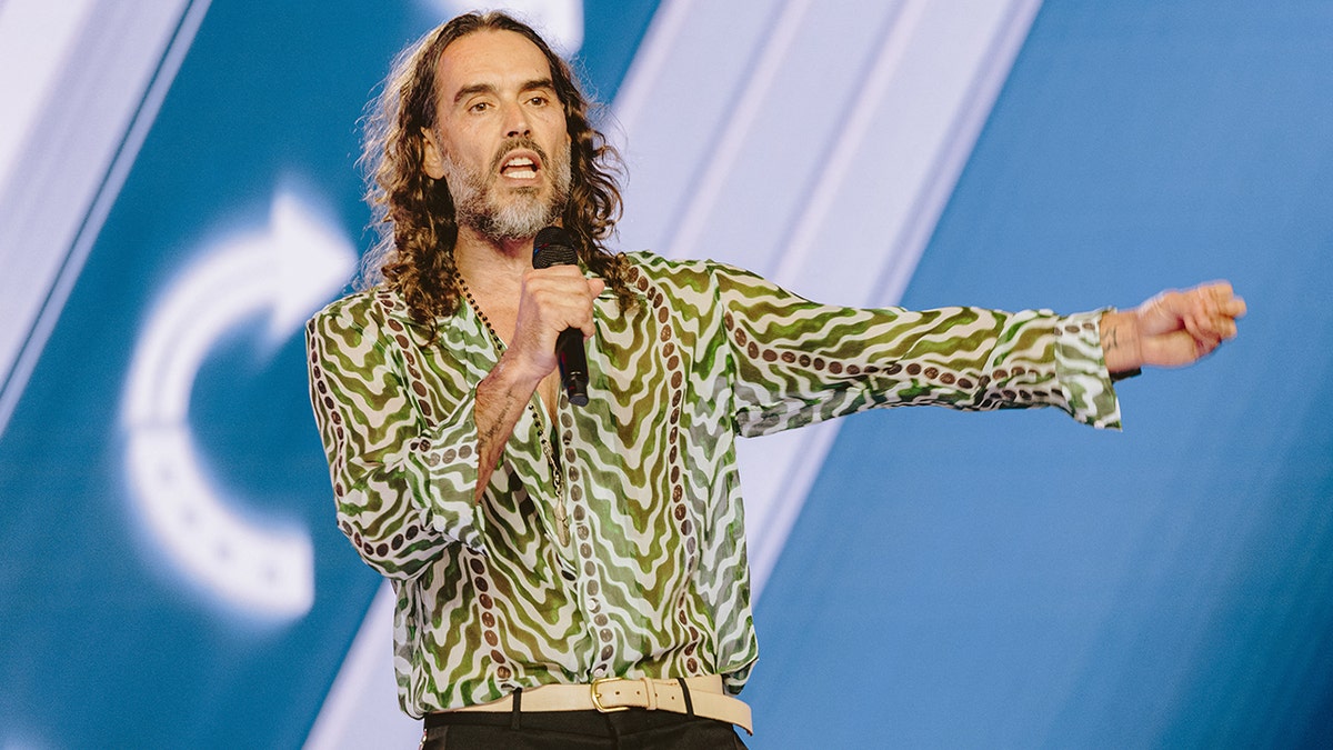 Russell Brand standing on stage wearing a dark jacket and shirt
