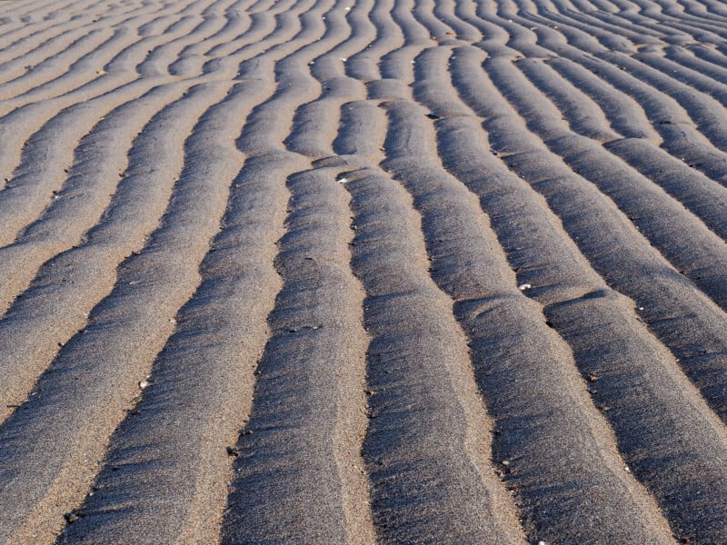 Close-up of smooth, wavy patterns formed in wet sand, likely by water or wind, creating parallel ridges and grooves that stretch into the distance. The lighting highlights the texture and contours of the sand.