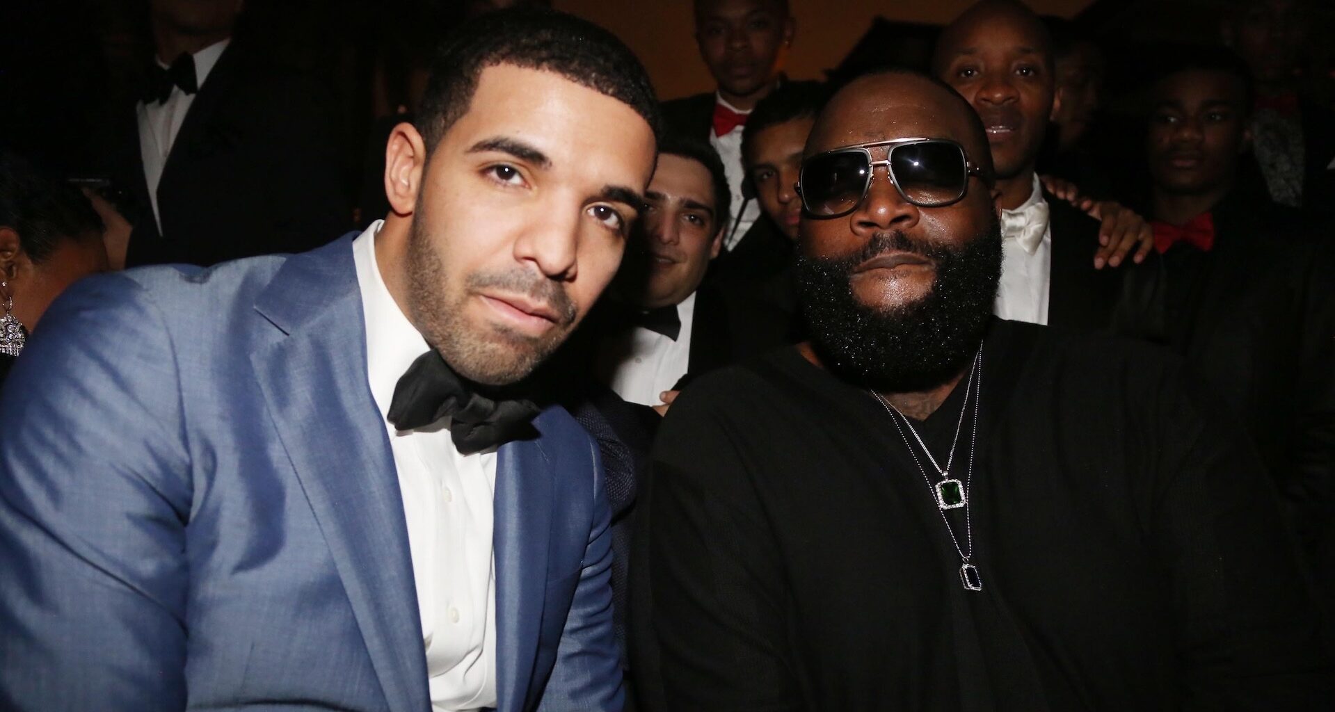 Drake in a blue suit and bow tie sits next to Rick Ross in sunglasses and a black shirt, surrounded by a group of people.
