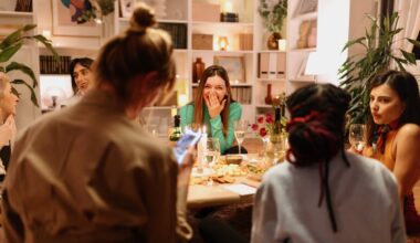 A group of women sit around a table with drinks and snacks, sharing money tips and laughing in a cozy, well-lit room with shelves and plants in the background.