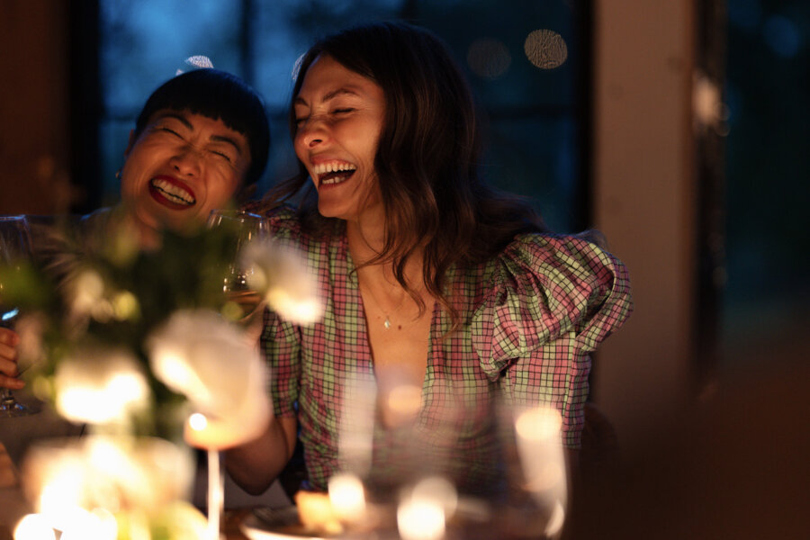 5 Things I Wish I Knew About Money in My 30s - 4 Two women sit close together at a dinner table, smiling and laughing as they share personal finance stories, with soft lighting and blurred flowers in the foreground.