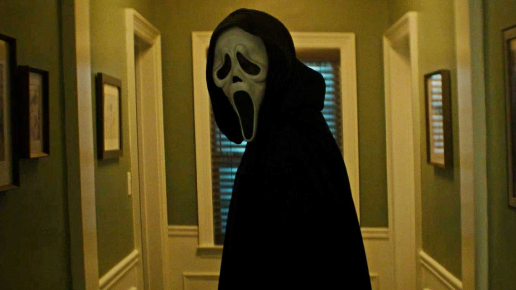 A figure in a hood and a Ghostface mask stares down a hallway, center framed, in a scene from Scream 7