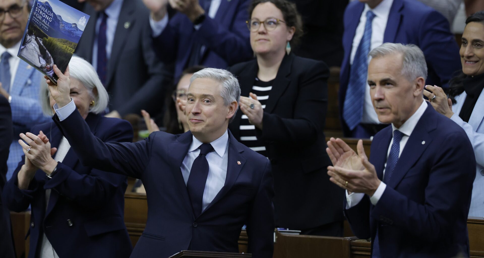 François-Philippe Champagne holds up a book while Mark Carney and other MPs applaud in the House of Commons.