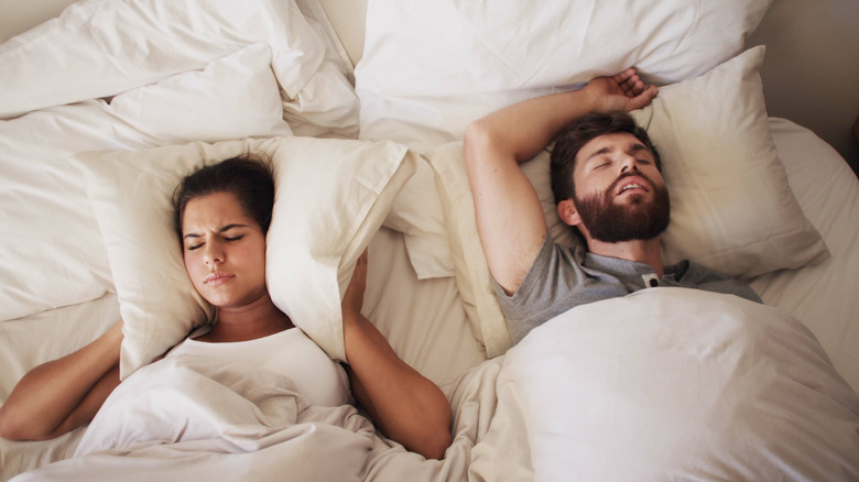 A man and a woman laying in bed together with the women holding a pillow over her ears while the man snores