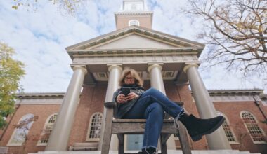 A woman sits legs crossed in a chair in front of a large university building shot from an extreme low angle.