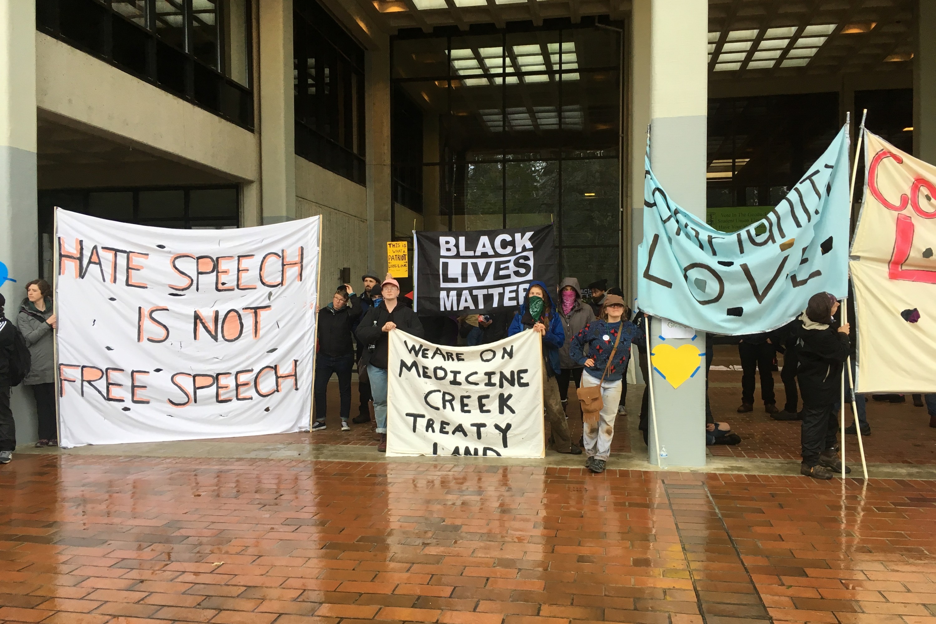 Inside an atrium, people hold signs reading 'Hate speech is not free speech', 'Black lives matter', 'We are on Medicine Creek treaty land' and others.