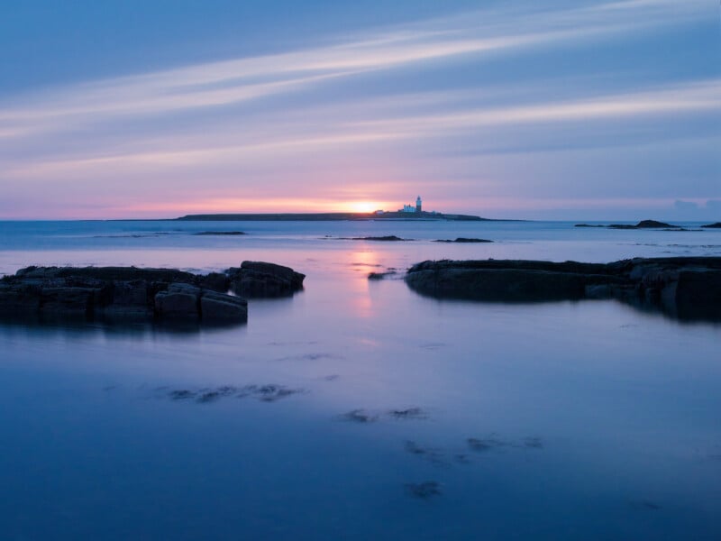 Calm sea at sunset with smooth water, dark rocks in the foreground, and a small island with a lighthouse and buildings in the distance. The sky is streaked with clouds and soft pink and purple hues.