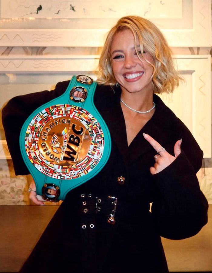 Young woman smiling and pointing at a colorful WBC championship belt in an indoor setting, highlighting influencer’s lie exposure.