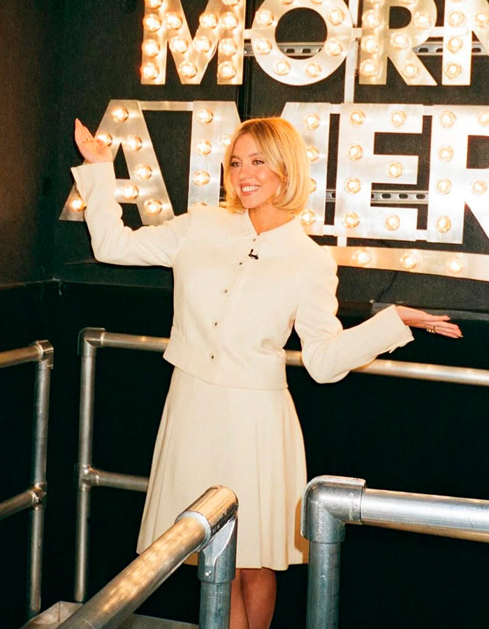 Sydney Sweeney smiling and posing with arms raised in front of a lit More at Eleven sign indoors.