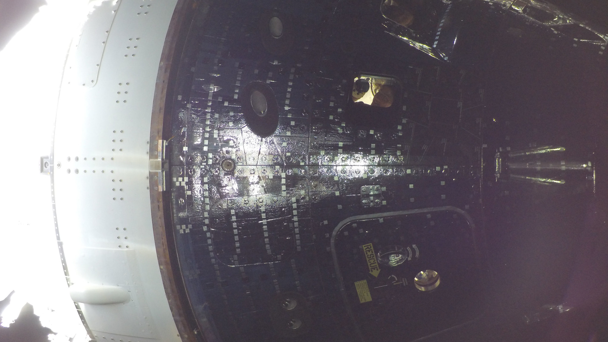 An astronaut can be seen smiling in the window of a dark spacecraft capsule as light reflects off its outer hull.