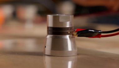An ultrasonic transducer with two wires attached to it by alligator clips floats very slightly suspended over a glass surface.