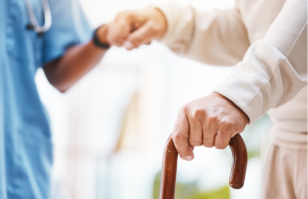 A person's hand with a walking stick in the foreground, with another person's hands clasped in the background.