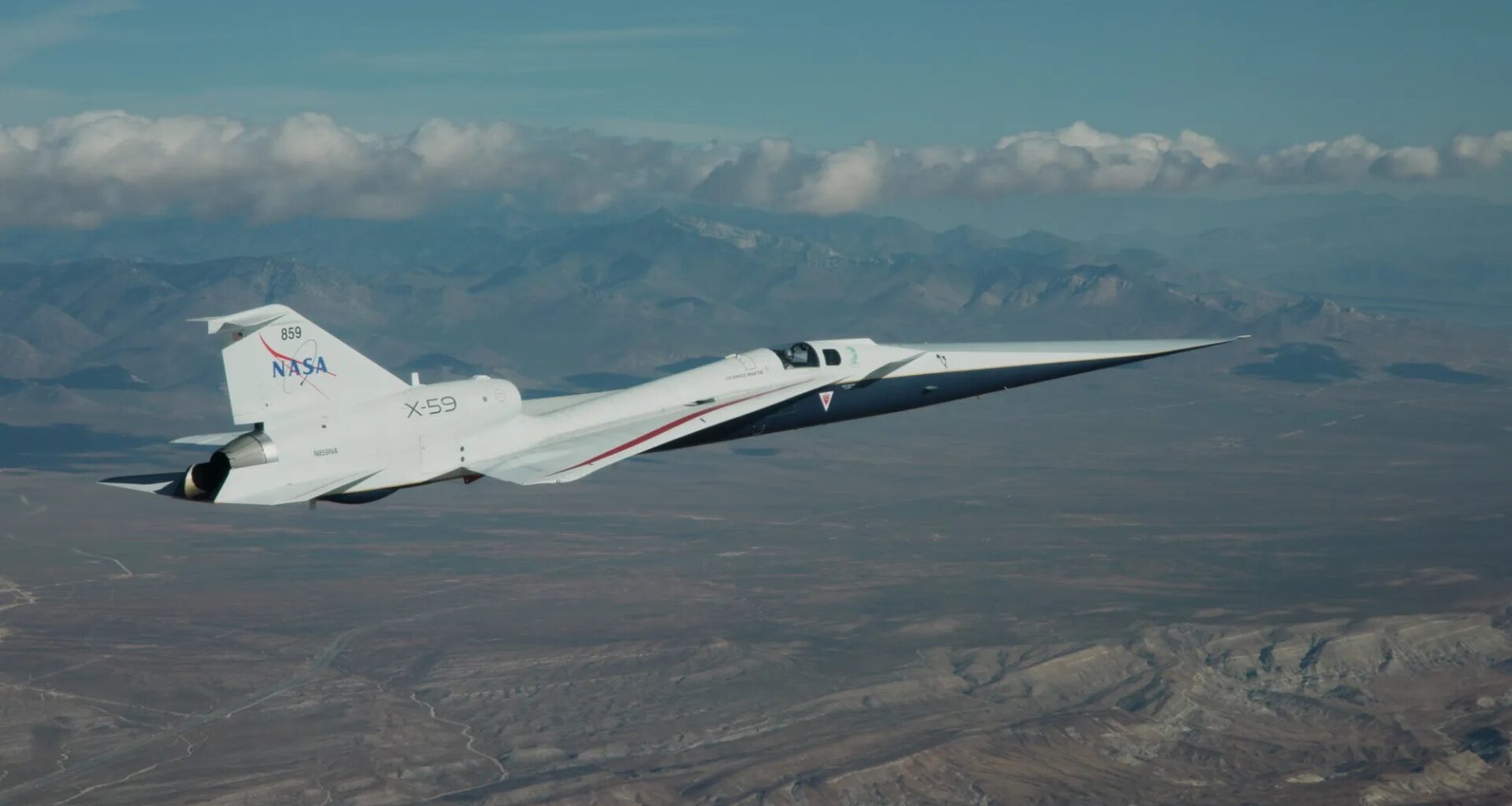 NASA’s X-59 flies above the Mojave Desert on a clear day. The white aircraft has light gray, red, and blue accents, with a NASA logo and the number 859 on its tail. It appears flying level over the desert landscape, with a mountain range visible on the horizon and a trail of clouds above.