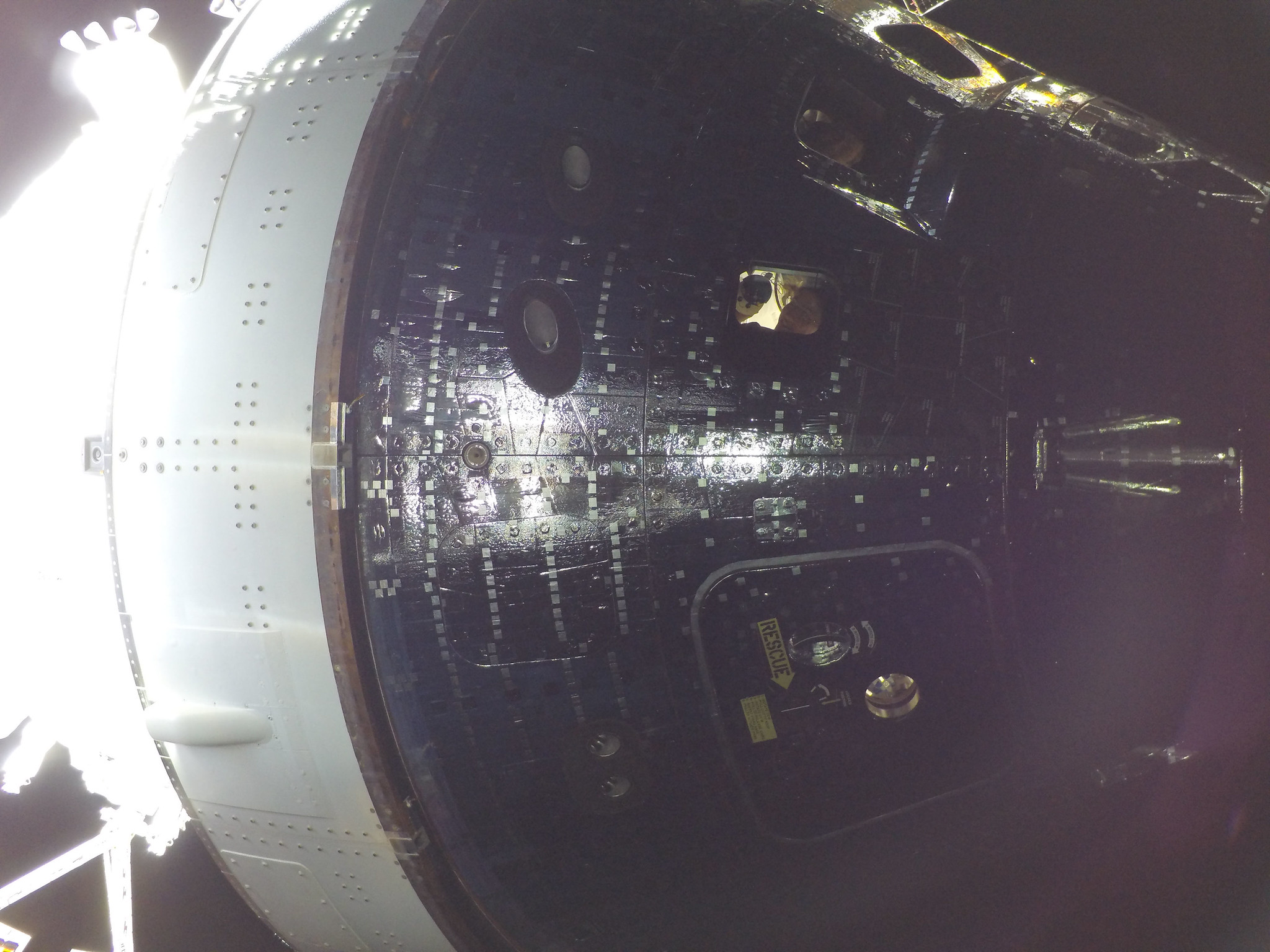 An astronaut can be seen smiling in the window of a dark spacecraft capsule as light reflects off its outer hull.