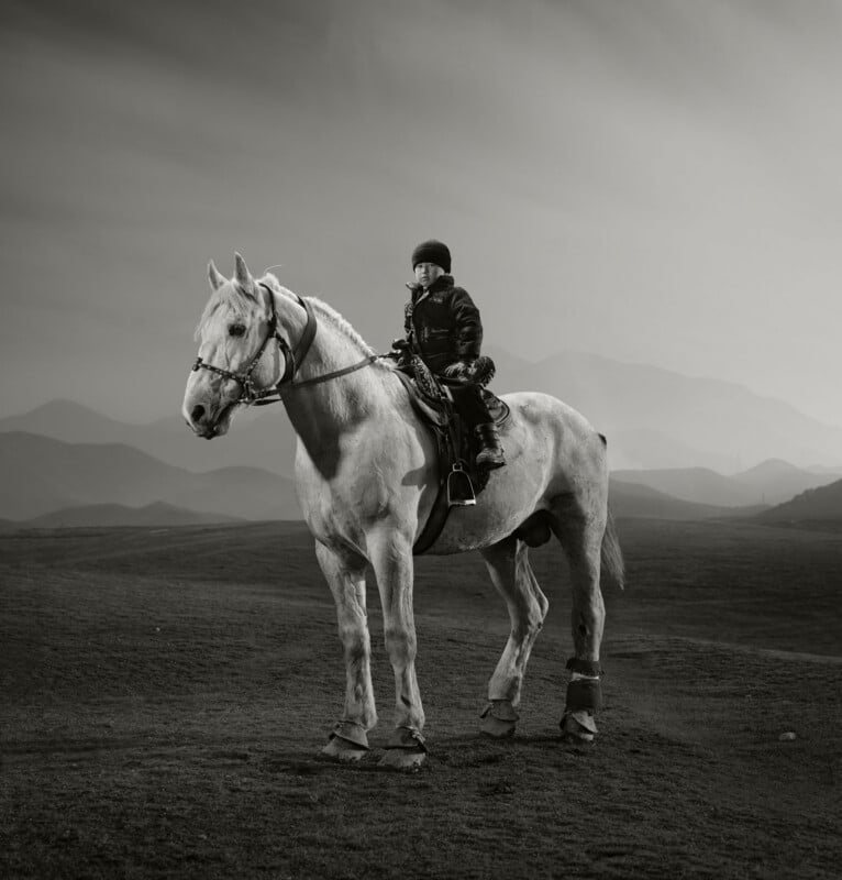 A person wearing a helmet and jacket sits on a saddled horse in an open landscape with distant mountains under a cloudy sky. The image is in black and white.