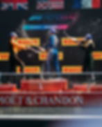 Three racers spray champagne on podium, celebrating at F1 Academy, Montreal. Flags and Pirelli signs in background, bright day mood.