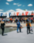 Racers in colorful suits walk on a dock holding helmets. Crowd watches in front of banners with racers' photos and numbers, under clear sky.