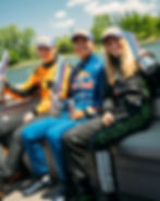 Three smiling race car drivers in colorful suits and caps hold trophies on a boat by a lake. Sunny day with trees in the background.