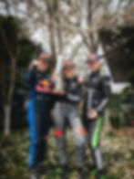 Three race car drivers in colorful suits and Pirelli caps celebrate with trophies under a blooming tree, smiling in a joyful outdoor setting.