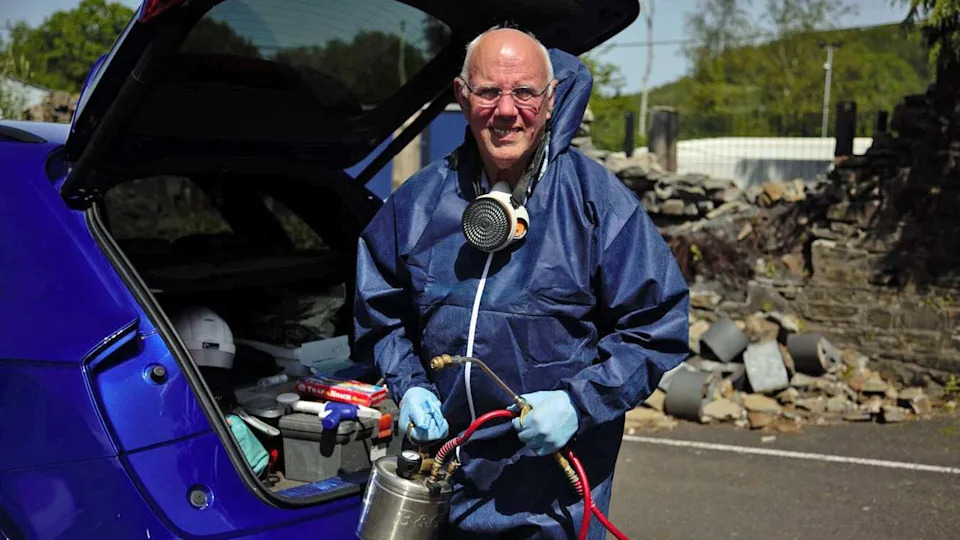 A man with white hair and glasses is wearing a blue body suit and blue plastic gloves, whilst holding a fumigating device