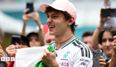 Kimi Antonelli celebrates his race win in Miami while holding the Italian flag. A crowd are gathered around him and taking photographs