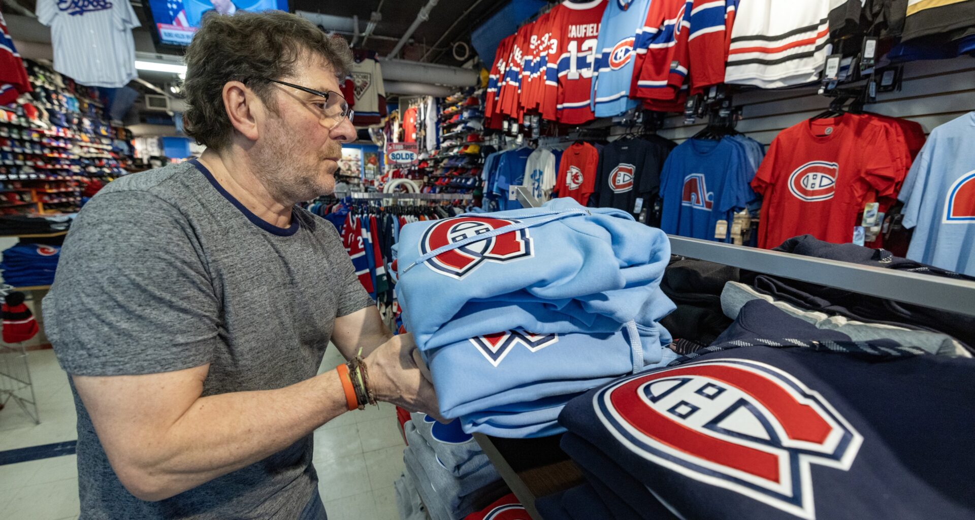Tony Araujo holds a pile of light blue Canadiens hoodies above a shelf in a sports apparel store.