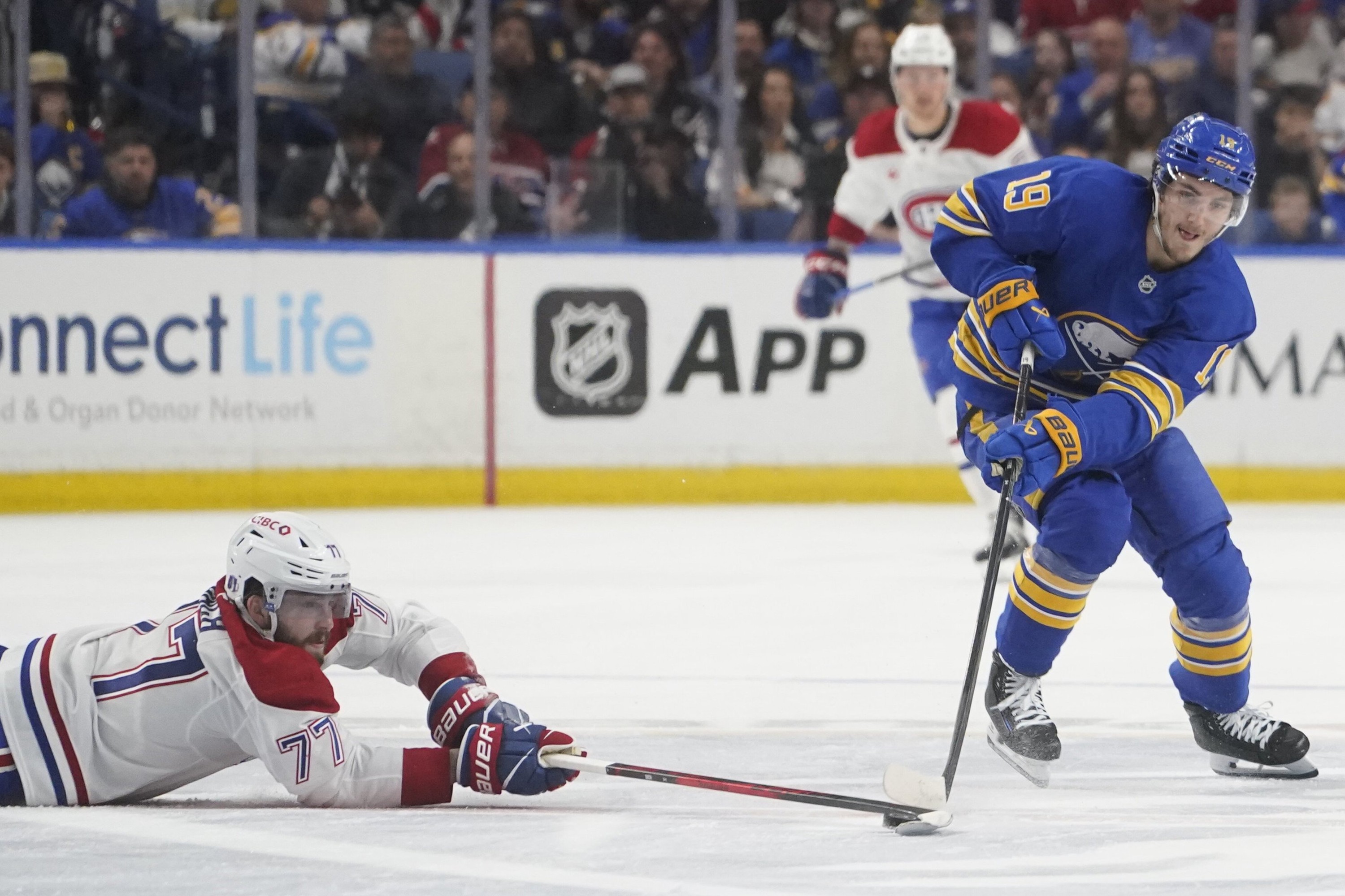 Buffalo Sabres centre Peyton Krebs (19) battles for a loose puck with Montreal Canadiens centre Kirby Dach, who is lying on the ice and reaching with his stick.