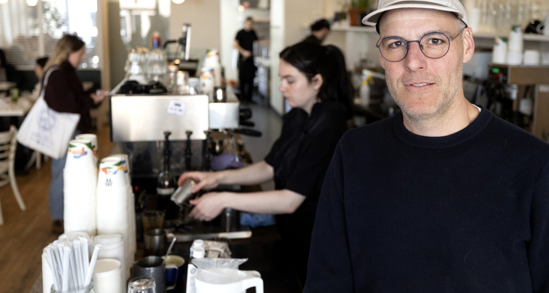 Simon Defoy stands in a café with staff preparing orders in the background.