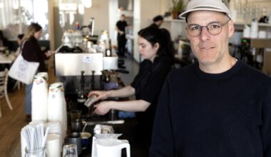 Simon Defoy stands in a café with staff preparing orders in the background.
