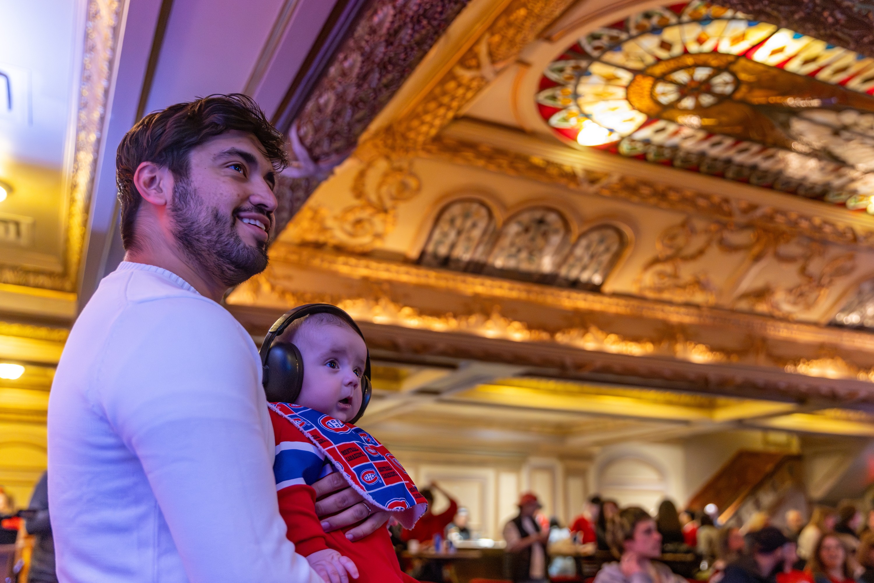 A man holds a baby wearing a Habs jersey and headphones in a theatre